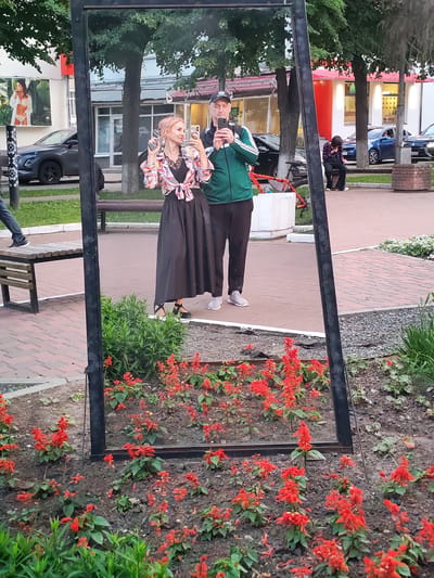 The image shows a full-length mirror placed outdoors, reflecting a street scene in Yoshkar-Ola, Russia. In the reflection, a couple is taking a selfie. The woman, with blonde hair, is wearing a black dress with a colorful tied-front blouse and holding her phone. The man, wearing a green tracksuit and a baseball cap, is also holding a phone. They appear to be on a paved walkway, with a park bench and greenery visible behind them. In the background, there are buildings with shops, trees, and cars. A red bicycle is leaning against a white planter. The time of day appears to be late afternoon or early evening, given the lighting. The overall mood is casual and relaxed. There is no visible text in the image.