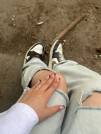 This high-angle, close-up shot captures a person from the waist down, seated or crouching in an outdoor setting in Novi Sad, Serbia. The subject wears light-wash, ripped denim jeans with visible skin through the tears, and sneakers featuring black, white, and a dark olive green swoosh. One hand rests casually on their knee, showcasing light pink nails with black French tips and a simple silver ring on the ring finger. A white long-sleeved garment is partially visible on the arm.
The setting is a natural, unpaved ground, likely a park or wooded area, covered in dry dirt, scattered pine needles, and small organic debris. Notable objects on the ground include a long wooden stick positioned diagonally near the left foot, a visible pine cone in the upper left corner, another partially seen pine cone in the upper right, and a discarded cigarette butt near the center-left. The lighting suggests it is daytime, possibly under an overcast sky, providing soft, diffused light. There is no discernible activity beyond a moment of rest. No text is visible in the image.