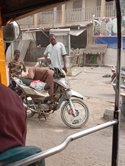 In Gawuna, Nigeria, a daytime street scene is captured from within a vehicle, likely an auto-rickshaw, evidenced by the yellow frame, silver handlebar, and maroon-clad passenger in the foreground. On a dirt road, a light-colored underbone motorcycle with missing body panels, exposing its engine, is centrally positioned. A patterned fabric is draped over its seat. A person in brown clothing is bent over the motorcycle's left side, appearing to inspect or work on it. To the right of this individual, a man in a light grey Henley shirt and dark trousers stands with his left hand on his hip, observing the scene with a neutral expression. To the left of the man, a boy in a camouflage shirt and dark trousers stands, holding a light-colored bottle in his right hand, looking downwards.
The background features a two-story building with balconies and white railings. Below it, several makeshift market stalls are constructed from wood and corrugated metal, with a blue tarp visible. One stall displays a large banner with images of food items. Scattered debris is present on the uneven ground. A second motorcycle is partially visible on the far right.