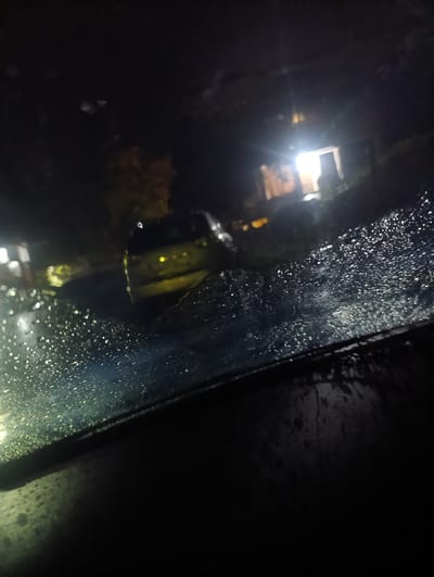 The image depicts a dimly lit outdoor scene at night, viewed through a wet windshield. Raindrops are visible on the glass in the foreground, distorting the view of the background. In the mid-ground, a yellow car is parked on what appears to be a road or driveway. Behind the car, a building with illuminated windows is visible, suggesting a residential area. The overall ambiance is dark and moody, consistent with a rainy night. The location is identified as the city of Gagra, Abkhazia. There are no people visible in the image.