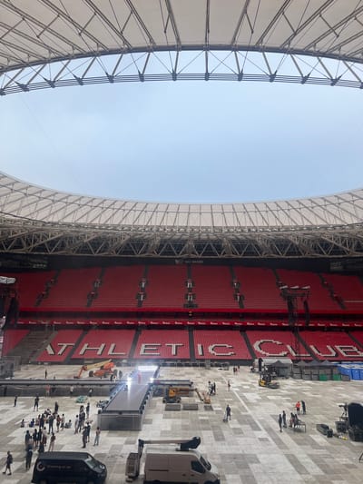The image shows a wide, low-angle view of the interior of a stadium with a retractable roof structure overhead. The stadium has red seating sections that wrap around a large, flat, concrete floor. Large white letters are visible on the seating sections, spelling out "ATHLETIC CLUB".

On the stadium floor, there are several construction and event preparation elements. A raised black platform runs across a portion of the field, with a person in a red outfit standing on it. There are several vehicles, including a scissor lift, a forklift, and two vans. A group of people are scattered across the floor, some appearing to be workers and others spectators or participants. Some are gathered in groups, while others are walking or working individually.

In the background, behind the red seating, there are darker sections that appear to be infrastructure or screens. The sky visible through the open roof is overcast. The scene suggests preparation for an event, possibly a concert or a sports ceremony, taking place in the San Mamés stadium in Bilbao, Spain.