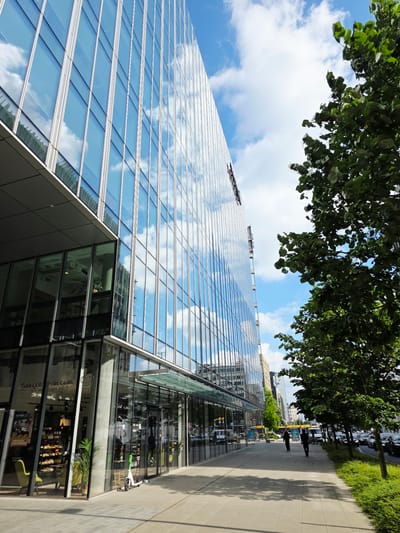This is a street-level, eye-level shot looking up at a modern, multi-story glass-fronted office building in Warsaw, Poland, likely in a business district. The building's facade is highly reflective, mirroring the blue sky and scattered white clouds. A mature tree with lush green foliage grows on the right side of the frame, partially obscuring the building and the sky.

In the foreground, a wide paved sidewalk runs along the building's base. The sidewalk is made of light-colored square tiles. On the sidewalk, in front of the building's ground-floor storefronts, are two people walking away from the camera, dressed in dark clothing. To the left of the sidewalk is a bright green electric scooter parked upright.

The ground floor of the building features large glass windows, offering a view into a shop with displays of baked goods and other items. Inside, a yellow chair is visible near the window.

Further down the sidewalk, more trees line the street, and traffic, including cars and a yellow bus, is visible on the road. The scene suggests a sunny day in a bustling urban environment. The building's architecture is sleek and contemporary, characterized by clean lines and extensive use of glass.