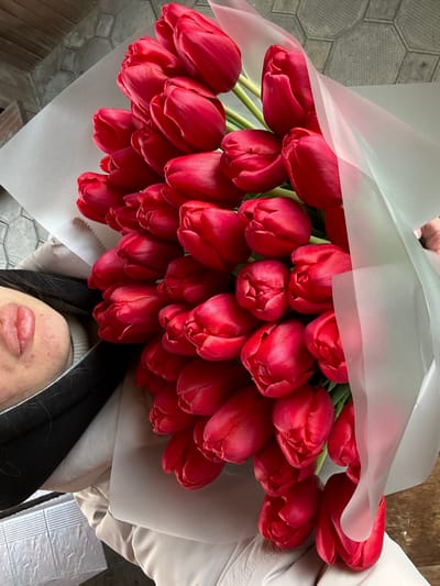 A close-up, top-down photograph displays a large bouquet of approximately 40-60 vibrant red tulips, tightly clustered and mostly in bud or partially open, wrapped in matte white paper and a translucent light-colored film. The bouquet is held by an individual, whose partial upper body is visible. On the left, a segment of the individual's left arm or shoulder is seen, covered by a black outer garment over a lighter, possibly ribbed, inner garment. A distinctive, raised, reddish scar or body modification, approximately 3-4 cm in length and consisting of two prominent lobes, is present on the visible skin of the arm/shoulder. The individual's lower torso or lap is covered by light-colored, possibly beige or tan, clothing. A hand is partially visible on the far right, supporting the bouquet. The background on the upper left features a grey, geometrically patterned surface, potentially paving or tiling. The background on the lower right is a darker, indistinct surface. The scene is located in Trudobelikovskiy, Russia.