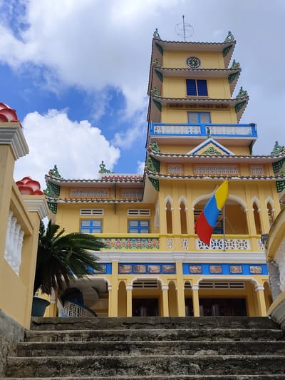 The image shows a close-up, low-angle view of a brightly painted yellow Buddhist temple or pagoda in Xuân Thành, Vietnam. The temple has multiple tiered roofs with ornate green and red trim. A flag with red, yellow, and blue horizontal stripes, likely a religious or cultural symbol, hangs from a balcony. The building features blue-trimmed windows and decorative friezes with colorful paintings. In the foreground, a set of weathered, stone steps leads up to the temple entrance. A palm tree is visible to the left of the steps. The sky above is a vibrant blue with scattered white clouds. The signage on the building appears to be in Vietnamese.