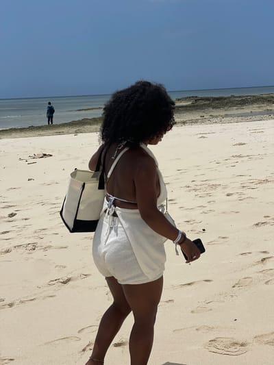 The image displays a bright, sunny beach scene in Zanzibar, Tanzania. A woman, observed from a rear-right perspective, is walking rightward on light-colored sand, leaving footprints. She has dark, curly hair and wears a cream-colored textured two-piece outfit, including a halter top with a tie-back and matching shorts. She carries a cream and black two-tone tote bag over her left shoulder. Her right hand holds a dark-colored smartphone, and she wears a white wristband and a brown beaded bracelet on her right wrist, with a thin chain visible on her left ankle. In the distant background, a person in a blue shirt stands in shallow ocean water, facing away from the camera. The midground water features dark, exposed coral or rock formations. The sky is clear and light blue, indicating fair weather.