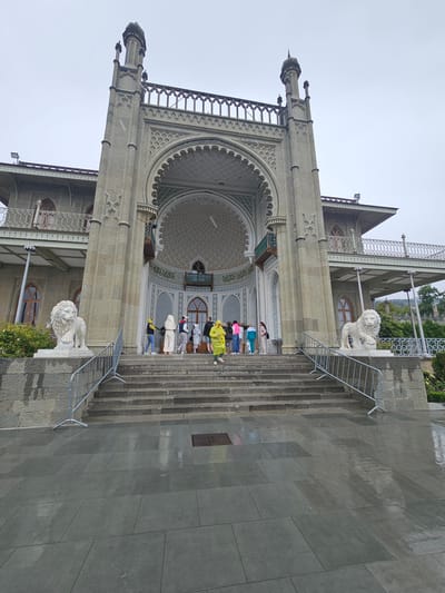 The image shows the entrance to a building with an ornate, Islamic-inspired design. The location is in the city of Алупкинский городской совет, Ukraine. A group of people are on the steps leading to the entrance. Two lion statues flank the stairs. The building is constructed of light-colored stone.