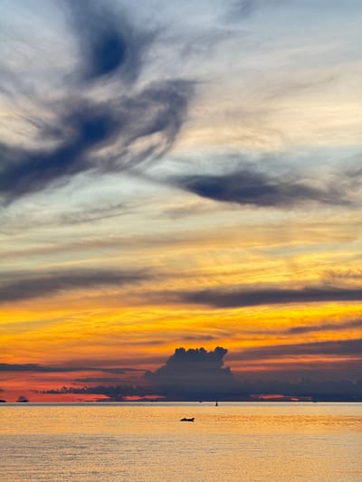 The image depicts a serene seascape at sunset on Koh Phangan, Thailand. The sky is a canvas of vibrant colors, transitioning from deep blues and grays at the top to fiery oranges and yellows near the horizon. Wispy clouds stretch across the sky, some appearing dark and dramatic, while others are soft and ethereal. The ocean is calm, reflecting the warm hues of the sunset. In the distance, small boats are visible, appearing as tiny silhouettes against the illuminated water. One boat is a dark, flat object in the foreground, while a buoy with a small flag stands further out. The overall impression is one of tranquility and natural beauty, characteristic of a tropical sunset.