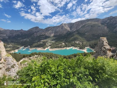 The image is a landscape view of El Castell de Guadalest, Spain. The foreground features vibrant green foliage partially obscuring a turquoise reservoir lake. In the distance, a range of mountains forms the backdrop. The sky is bright blue with scattered cumulus clouds. A rocky outcrop is visible on the right side of the frame, while a smaller stone structure appears to the left. The lake has an irregular shape with branching sections, indicative of a reservoir formed in a valley. The mountains are sparsely vegetated with trees.