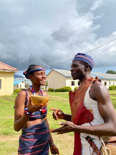 This media file captures a vibrant cultural moment in Wang, Nigeria. Two individuals, a man and a woman, are engaged in conversation outdoors. The man, wearing a white tank top, a traditional leather vest, and a blue and red striped cap, is looking down at the woman. The woman, adorned in a striking striped outfit and a matching headband, smiles warmly as she holds a halved gourd. Her ensemble is further enhanced by colorful beaded necklaces and bracelets.
The scene is set against a backdrop of grassy terrain and several buildings, suggesting a semi-urban or rural environment. The sky is overcast, with dramatic clouds hinting at impending rain. The lighting indicates it is likely daytime, possibly late afternoon given the soft light.
The interaction between the two individuals appears to be friendly and animated, conveyed through their body language and the woman's open smile. The presence of the gourd suggests a possible cultural or ritualistic element to their gathering, perhaps related to traditional crafts or food preparation. The overall impression is one of cultural pride and community interaction within a Nigerian setting. No visible text is present in the image.