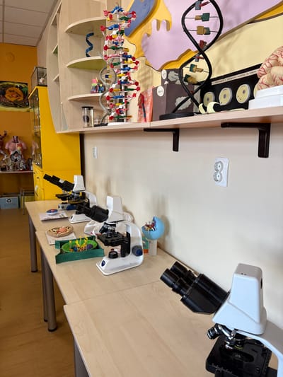 The image depicts a science lab or classroom setting, likely in Panagyurishte, Bulgaria, although this is only inferred from the image's potential origin. It features a light-colored wooden desk cluttered with science-related models and equipment. Three microscopes are visible, positioned at various angles. To the left, there is a bookshelf or display case containing anatomical models, including a brain, a DNA model, and diagrams illustrating human development. The overall impression is that of a learning environment focused on biology or life sciences. A recycling bin labeled "ХАРТИЯ" is also visible.