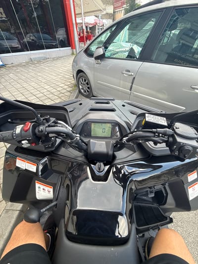 The image is a first-person view from a black ATV parked on a sidewalk in Gabrovo, Bulgaria. The ATV has a digital display showing "0" and "146" with speed and distance information. The handlebars have controls for lights and indicators, as well as a red button. A person's legs, clad in black shorts and black and white sneakers, are visible in the foreground. To the right of the ATV, a silver car is parked. In the background, reflections of buildings and trees are visible in the windows of a building. The sidewalk is paved with grey tiles. There are umbrellas and tables visible in the background, suggesting a cafe or restaurant setting. Two warning stickers are affixed to the left side of the ATV.