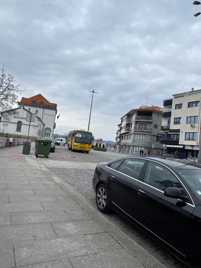 The image depicts an urban street scene in Bragança, Portugal, specifically near the former Bragança train station, identifiable by the "BRAGANCA" lettering on its white facade. The sky is overcast with light grey clouds.
In the immediate foreground on the right, a black four-door sedan, consistent with an Audi A6 model, is parked parallel to a grey stone slab sidewalk. The front of this vehicle is out of frame. The sidewalk extends across the lower left portion of the image.
Adjacent to the sidewalk, the street is paved with dark cobblestones, sloping slightly downwards towards the right. On the left side of the cobbled street, a dark green wheeled waste bin and a dark grey litter bin are positioned near the sidewalk. Behind these bins, a dark-colored car is partially visible.
Further along the cobbled street, a yellow bus is positioned, facing slightly towards the left. Behind the bus, a white commercial van is visible. In the middle-ground, a tall, dark grey street light pole stands.
On the left side of the scene, behind the waste bins, stands a multi-story white building with a prominent reddish-orange tiled roof and several windows. This structure is the former Bragança train station. A tree with bare branches is partially visible on the far left.
On the right side of the street, a sequence of multi-story buildings lines the sidewalk. One building features several balconies with grey railings, and another is predominantly white with blue accents around its windows. Two individuals are observed walking on the sidewalk in front of these buildings, moving away from the viewer. A red postbox is visible on the sidewalk further down on the right. Distant hills are faintly visible under the overcast sky in the background.