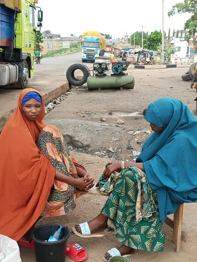 Two women, dressed in traditional Islamic attire, are seated on the ground in what appears to be a marketplace or roadside rest stop in Gadao, Nigeria. The woman on the left wears an orange hijab and a patterned skirt, while the woman on the right is adorned in a blue hijab and a green patterned skirt. Both women are facing inwards, engaged in conversation or a shared activity, their hands clasped in their laps.

The scene is a bustling environment, evidenced by the presence of large trucks, a motorcycle, and various pieces of equipment, including an air compressor and tires. The ground is dirt and gravel, typical of an unpaved area, with scattered debris suggesting a functional, albeit possibly untidy, public space. In the background, residential buildings and trees indicate a semi-urban or fringe-urban setting. A black bucket containing a bottle of water and a red flip-flop are visible near the woman on the left, while the woman on the right has a white slipper on her foot. The overall impression is one of everyday life unfolding in a vibrant, possibly commercial, district of Gadao.
