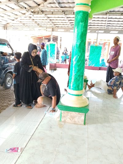 The image shows a scene in front of a building in Anjatan, Indonesia (latitude: -6.419, longitude: 107.979). A woman wearing a black abaya is talking to a man in a black t-shirt and brown shorts while another man sits to the left, observing. Both men are wearing head coverings. The woman's hand is on the man's arm.  A green pillar with a white base stands between them, partially obscured by the woman's long black abaya.  A second man in a blue shirt sits to the right, with his back to the camera, while a man in a pink shirt sits to the right with a white turban. Both men have their legs outstretched. The background includes a partial view of a white van. There is a large, partially open metal structure with a metal roof overhead. The ground is covered in white square tiles. The scene suggests a public place in Anjatan.