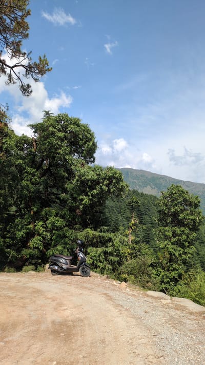 The image shows a scene in Dharamkot, India. A black scooter is parked on the side of a dirt and gravel road. Lush green trees and dense vegetation border the road, partially obscuring a mountain in the background. The sky is blue with scattered white clouds.
