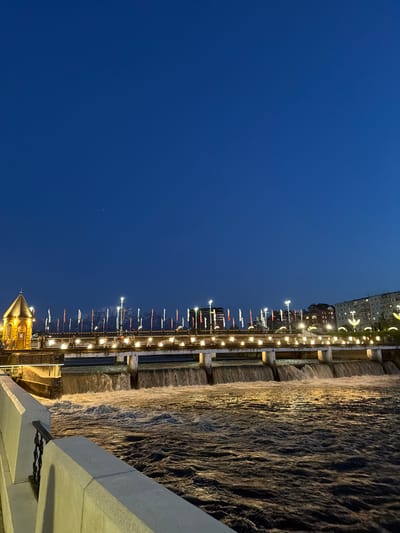 The image presents a nighttime scene in Chermen, Russia, featuring a river flowing beside an illuminated bridge. The sky above is a deep blue. The river's surface reflects the lights from the bridge, creating a shimmering effect. The bridge is lined with lampposts and what appear to be banners or flags. Alongside the river is a concrete barrier. Buildings can be seen further along.