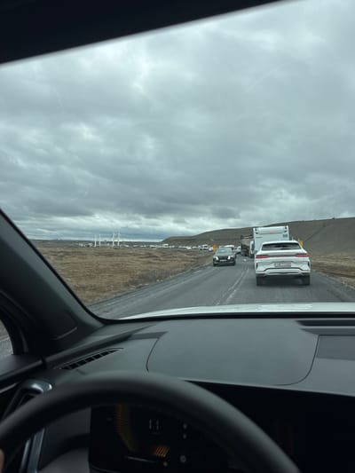 The image captures a road scene from the perspective of a vehicle's interior, looking forward through the windshield. A line of vehicles is visible ahead on a two-lane road. The most prominent vehicle is a white SUV, followed by a large white camper or truck, and several other cars further down the road. The white SUV has a license plate reading "HE U46".

The surrounding landscape features barren, brown hills under a heavily overcast, cloudy sky. In the distance, on the left side, there appear to be large, white structures, possibly power lines or part of a distant bridge or industrial facility.

Inside the vehicle, the steering wheel and a portion of the dashboard, including a digital display with illuminated elements, are visible on the right side of the frame. The left side of the frame shows the car's interior door panel and part of the windshield.

The activity taking place is driving, likely in a queue or convoy. The weather is overcast. Given the barren, volcanic-like landscape and the license plate format, the scene is consistent with Iceland. It is daytime, despite the cloudy conditions. No people are directly visible.
