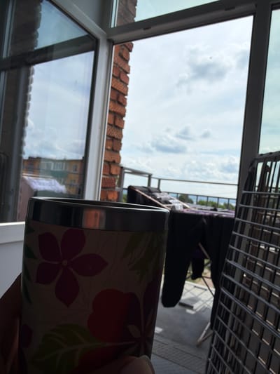 A close-up shot focuses on a person's hand holding a floral patterned mug in the foreground. Beyond the mug, a balcony view is visible through a large window. The balcony features a drying rack with dark clothing hanging on it, a black plastic chair, and a silver metal drying rack. In the background, a body of water is visible, likely the Kama River, with a distant shoreline. The sky is overcast with scattered clouds. The overall scene suggests a domestic setting on a balcony overlooking a natural landscape, possibly in Sarapul, Russia, given the reference to the Kama River.