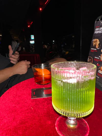 The image shows a low-light indoor scene, presumably in a bar or restaurant in Canggu, Indonesia, as evidenced by the menu item descriptions on the table. A person is holding a phone in their left hand. On the red velvet tablecloth are two cocktails: one green, tall, and ornate with pink elements and a stem, and another amber-colored drink in a smaller glass. There is a menu on the table.  In the background are lights suggesting a cityscape or busy street, with the logo of 7-Eleven visible.