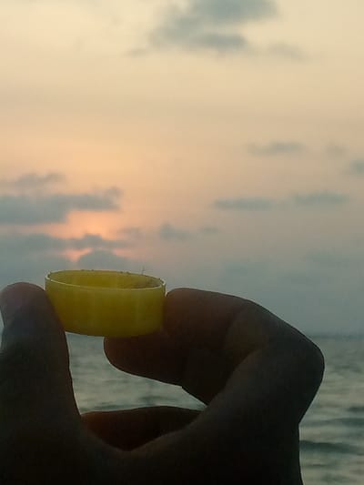 A hand holds a yellow plastic bottle cap up against a cloudy sky at sunset. The sun is obscured by the clouds. Below, the sea stretches out into the horizon, with calm waves reflecting the sky's soft colors. The bottle cap is the central focus, held between the thumb and forefinger. The overall tone is serene and contemplative.