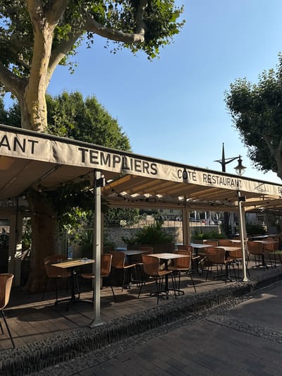 This is an outdoor scene of a restaurant's seating area. The image shows a row of tables and chairs set up on a cobblestone patio, shaded by a large awning. The awning is a light beige color and has text printed on it. The tables are small and square, made of a light-colored wood, and are set with placemats. The chairs are made of woven wicker with black metal legs.

There are no people visible in the image, but the setup suggests the restaurant is open or preparing to open. The time of day appears to be daytime, likely late morning or early afternoon, given the bright sunlight casting shadows. The weather seems pleasant and sunny.

The awning has large, dark text that reads "TEMPLIERS COTE RESTAURANT". Behind the awning, greenery from trees and plants is visible, suggesting a location with a moderate climate. The cobblestone pavement and the style of the restaurant facade in the background hint at a European setting, specifically Collioure, France, as indicated by the location context. The overall impression is one of a charming and inviting outdoor dining space.