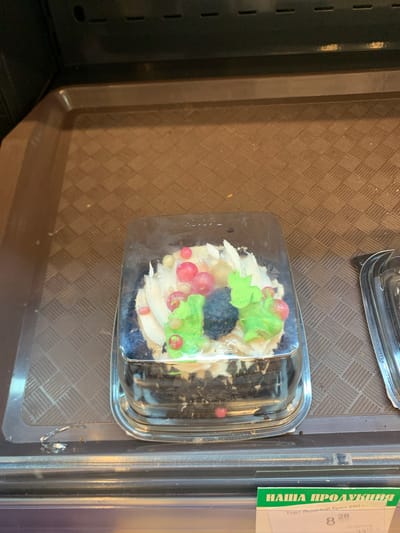The image shows a single, individual-sized cake in a clear plastic container, placed on a brown textured shelf within a retail environment. The cake is decorated with white frosting, pink and green confectionary berries, and a dark, possibly chocolate, element resembling a blackberry or truffle. The background consists of a dark, ribbed surface, characteristic of a display shelf in a store. A green label with white lettering is partially visible at the bottom of the image, suggesting this is likely from a supermarket or bakery. The overall scene is one of a commercial product display. The location is specified as Kapyĺ, Belarus.