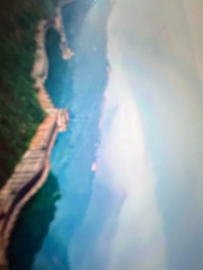 The image is a highly blurred, aerial view of a serpentine structure, likely a wall or road, winding through a landscape of green vegetation and a body of water. The structure appears to be made of stone or concrete and has a distinct, segmented pattern. To the right of the structure, there is a vast, bright, and hazy area, possibly indicating fog or clouds. The overall impression is that of a natural landscape with a man-made element cutting through it.

The image does not contain any discernible human figures, specific city landmarks, or identifiable features that would allow for pinpointing its location in Gelendzhik, Russia. The extreme blurriness obscures any details that could assist in geographic identification.