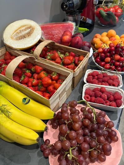 A still life image displays a variety of fresh fruits arranged on a dark countertop, likely in a kitchen setting. Prominently featured are two wooden crates overflowing with bright red strawberries. To the left, a halved honeydew melon, wrapped in plastic, rests on one of the crates. In front of the crates, a bunch of three yellow bananas, one with a "Yellow Guatemala" sticker, lie horizontally. To the right of the strawberries, two white cardboard punnets are filled with vibrant red raspberries. Above the raspberries, a white bowl is packed with dark red cherries. Further to the right, a pile of apricots and peaches are visible, along with a slice of watermelon. In the background, a red juicer machine and a glass bowl containing tomatoes and cucumbers can be seen. Red grapes are arranged on a pink heart-shaped plate in the foreground, partially obscuring the bananas. The lighting suggests an indoor environment, possibly a domestic kitchen, with a neutral grey background. The overall scene is a colorful and abundant display of fresh produce.