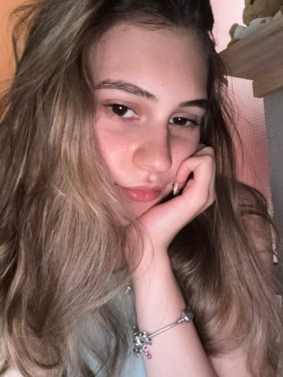 This is a close-up, horizontally oriented, selfie-style photograph of a young person, likely a female, with long, wavy, light brown hair. The subject is lying down, with her head tilted and her hair spread around her face. Her right hand is resting on her cheek, and her left arm is visible, adorned with a silver bracelet featuring charms, including one pink charm.
The subject's facial features are visible: her left eye, eyebrow, nose, and mouth. Her expression appears relaxed and contemplative. The lighting suggests it might be indoors, possibly during the daytime, given the natural light illuminating her face. The background is out of focus, but hints of a light pink textured surface and a wooden object suggest an indoor setting, possibly a bedroom. There is no visible text in the image. The overall mood is intimate and candid.