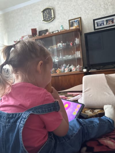 In Verkhnerussky, Russia, a young girl is lying on a couch, engrossed in an activity on a tablet. She is wearing a pink t-shirt and blue denim overalls, with her hair tied in pigtails. The tablet displays a purple screen with colorful icons and a large flower, suggesting an interactive game or educational app. The background reveals a living room setting, featuring a glass-fronted cabinet filled with decorative items, a large television screen, and framed pictures on the wall. A wall clock with a black face and gold numbers indicates the time. The overall mood is one of quiet concentration and domestic comfort, with no indication of the time of day or weather outside.