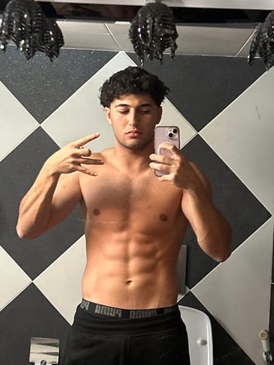 An image taken in Sarouja Municipality, Syria, depicts a young adult male capturing a mirror selfie indoors. The individual, shirtless, with a muscular build and dark curly hair, stands facing a mirror. He holds a light-colored smartphone with a triple camera array in his right hand, extended to photograph his reflection. His left hand is raised, forming a gesture with the thumb and pinky extended while the middle fingers are curled. He wears black sweatpants with a visible grey "PUMA" branded waistband. The background features a wall covered in large, diagonally arranged black and white checkered tiles. Two dark, textured, possibly sequined, decorative fixtures hang from the ceiling above the subject. A portion of a white porcelain fixture, likely a sink or toilet, is visible in the lower right, and a metallic fixture is partially seen in the lower left.