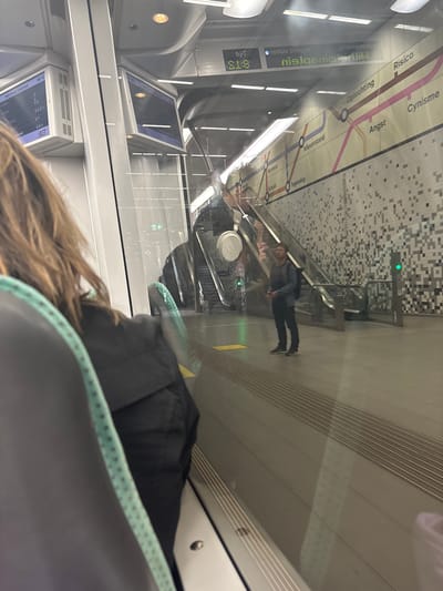 The image is taken from inside a train in the city of Rotterdam, Netherlands. It shows a reflection on the train window, looking out onto a metro station platform. In the reflection, there are two digital display screens above the platform, showing train information. The platform itself is visible with a wall adorned with a mosaic-like pattern of grey and white squares. Escalators are seen on the platform. A man in a dark jacket and blue shirt is standing on the platform, holding a phone. The train interior is partially visible in the foreground, with seats and handrails. The overall scene suggests the train is at a station or passing through one.