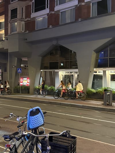 The image depicts a street scene at night in Amsterdam, Netherlands, with several people riding bicycles on a paved road that features tram tracks. In the background, a large, modern building with a distinctive architectural design is visible, featuring a grey, angular overhang supported by concrete pillars. The building has large windows, some of which are illuminated, and a brick facade on the upper levels. Red and yellow lights from a sign are visible in the distance, possibly indicating a business. Several bicycles are parked in the foreground, one of which has a blue child seat. The overall atmosphere is urban and nocturnal.