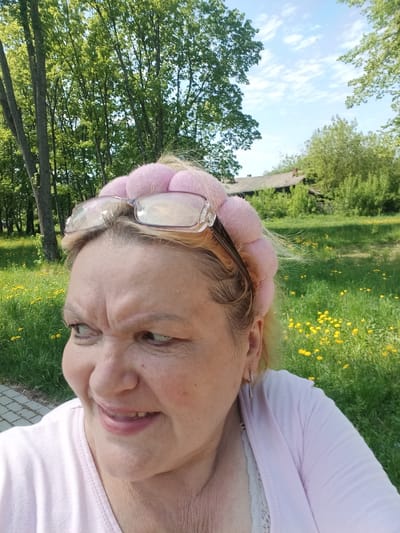 A close-up shot shows a woman in Красный Маяк, Russia, with a pink padded headband and eyeglasses perched on top. She wears a light pink top with a lace trim visible underneath. The background features a grassy area with yellow flowers, trees, and a building in the distance.
