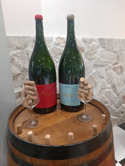 The image depicts a wine tasting setup on a wooden barrel, possibly within the city of Dub, Montenegro. Two large, dark green wine bottles with labels stand upright on the barrel's surface. The bottle on the left has a red cap and a red label that reads "Plantaze Stari podrum". The bottle on the right has a gold cap and a light blue label that reads "Plantaze Nota 2022".

In front of each bottle is a wine glass filled with corks. The glass in front of the left bottle contains an assortment of corks, some of which have visible inscriptions. The glass in front of the right bottle also contains corks, with some text visible on them.

Scattered on the surface of the wooden barrel are several individual corks, appearing to be for decoration or display. The barrel itself has dark metal bands and a textured wooden surface.

Behind the barrel, a wall constructed of natural stone blocks serves as the backdrop. The lighting in the scene is soft, casting a gentle illumination on the objects. The floor visible at the bottom right of the image appears to be tiled in a black and white checkerboard pattern. There are no people visible in the image.