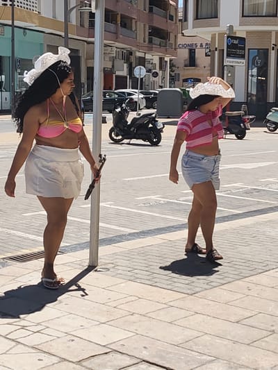 Two women are walking on a paved sidewalk in Dénia, Spain. The woman on the left wears a pink and gold bikini top, a white mini skirt, and white flip-flops. She also has a large white hat and is holding a black selfie stick. The woman on the right wears a pink and white striped crop top, blue denim shorts, and brown sandals. She is also wearing a large white hat and has her right hand holding it to her head. They are both walking away from the camera. In the background, there is a busy street with parked cars and motorbikes, as well as buildings with shops and apartments. A blue sign for "Raul Rubio FISIOTERAPIA" is visible.