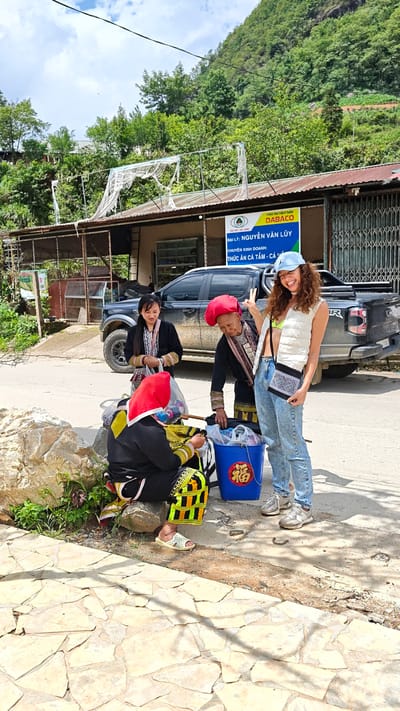 The image captures a scene in Sa Pa Ward, Vietnam, showing four women. Three are dressed in traditional clothing, including red headwear and black attire with colorful embroidered accents, seated near a blue bucket with a Chinese character. One is sitting on a rock on the side of the road while the others are standing next to her. The fourth woman, wearing a white vest, light-wash jeans, and a blue baseball cap, stands and smiles, holding up a peace sign.
Behind them, there is a black pickup truck parked in front of a single-story building with a metal roof and a sign reading "DABACO NGUYỄN VĂN LŨY," indicating a business specializing in aquatic animal food. The ground is a mix of stone pavement and asphalt. The scene is set against a backdrop of lush green hills and trees. The light is bright, suggesting it is midday on a clear day.