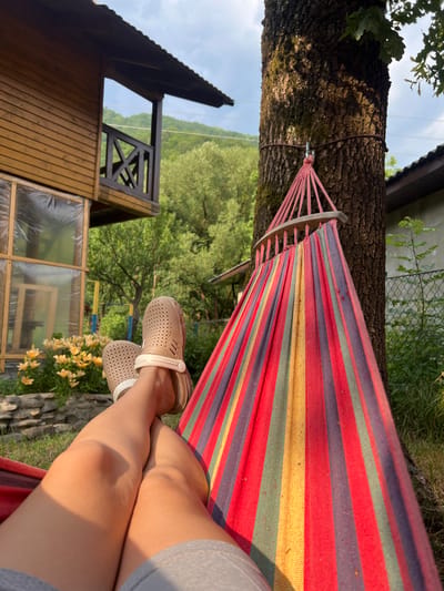 A person is relaxing in a colorful striped hammock slung between a tree and an unseen anchor point. The hammock has red, blue, yellow, and green stripes. The person is wearing light-colored clogs and their legs are visible, suggesting a warm day.
The scene appears to be a rural or natural setting, possibly a backyard or campsite. In the background, there is a wooden structure resembling a house or cabin with a balcony. Beyond that, lush green trees and hills are visible, indicating a mountainous or hilly landscape. The lighting suggests it is late afternoon or early evening, with the sun casting long shadows and highlighting the textures of the tree bark and the hammock. The weather seems pleasant, suitable for outdoor leisure. The overall impression is one of peaceful relaxation in a serene natural environment. The provided location context of Ust-Chorna, Ukraine, further grounds the scene in a specific geographical area known for its natural beauty.