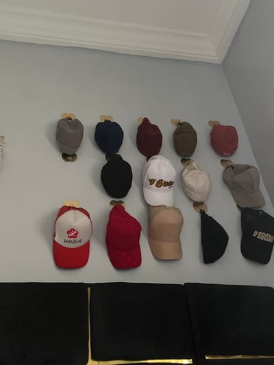 The image shows a collection of baseball caps mounted on a light gray wall. The caps are arranged in two columns and attached to the wall with small, round wooden pegs. The caps vary in color and style. In the background, there is a set of black cushioned panels with gold vertical stripes. The image does not provide any information to suggest a location within Kubwa, Nigeria.
