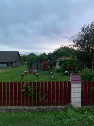 This image depicts a rural scene in Відзаўскі сельскі Савет, Belarus, likely a residential backyard. In the foreground, a dark red, wooden picket fence runs horizontally across the frame. To the right of the fence is a brick pillar topped with a burgundy roof, serving as a gatepost. Beyond the fence, a lush green lawn stretches towards the background.
To the left of the lawn, a weathered, dark-colored wooden barn with a corrugated roof is visible. In the middle ground, a variety of elements are scattered across the yard. A black trampoline with a safety net stands prominently. In front of it, an arched metal structure holds several potted flowers in shades of pink and white. Further back, a small, white gazebo with a burgundy roof is surrounded by dense green foliage and flowering plants. A red tree with dense foliage is also present in this area. The background is filled with mature trees and dense vegetation under a cloudy sky. The overall atmosphere suggests a tranquil, rural setting.