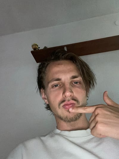 A young man with medium-length brown hair, a mustache, and a goatee is pictured from the chest up, looking into the camera. His right index finger is touching his lips. He is wearing a white waffle-knit shirt. Behind him, a dark brown shelf is mounted on a light grey wall. A small, yellow plush toy with a distressed expression sits on the left end of the shelf. The image appears to be taken indoors, possibly in a home, in Šušanj, Montenegro.