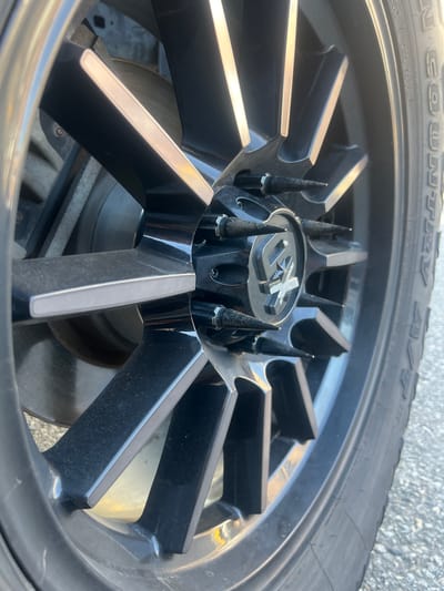 A close-up shot captures a black custom wheel with silver accents, featuring a spiky lug nut cover. The tire tread is visible on the right side of the image, with part of a brake rotor and suspension components showing behind the wheel. The scene appears to be outdoors, with an asphalt surface and gravel in the background, suggesting a street or parking lot environment. The lighting indicates it is daytime, possibly in the city of Richmond, Canada, given the context.