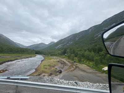 An overcast day in an unnamed valley in Norway. A wide, shallow river flows from left to right across the frame. The river is surrounded by a gravelly bank and then a lush green landscape of trees and rolling hills. In the foreground, a metal guardrail runs along a road. The right side of the image shows a reflection in a truck's side mirror, revealing more of the road and the imposing, tree-covered mountains. A small, overturned boat rests on the riverbank. The overall impression is one of natural beauty and rugged wilderness.