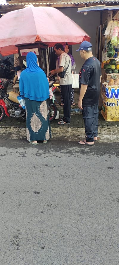 A street scene in Kecamatan Lawang, Indonesia (latitude: -7.847, longitude: 112.7), captured from a low angle. Four individuals stand on a paved street, three are facing towards a small food stand, one is standing slightly behind the group, facing away from the stand. The food stand is beneath a pink umbrella, with various items and products displayed. The first individual in line is wearing a light blue hijab and green and white dress, standing in front of the stand. The second individual in line is a man wearing a grey t-shirt, dark pants, and carrying a black bag across his body. He is looking down at the stand. Behind the second individual is a woman with long, black hair wearing a bright pink and purple scarf. She is facing the stand. The last individual in line is a man wearing a black shirt and black pants. His head is turned and facing the camera. All four individuals are standing on a gray pavement, which extends out from the stand. The background is slightly out of focus, but shows a wall with a visible sign with text that reads "AWAN klatan").