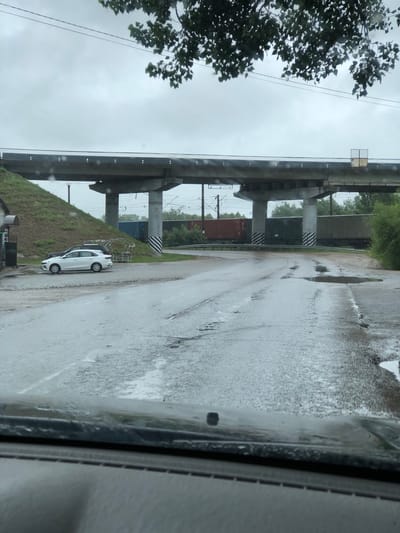 The image shows a wet, grey day under an overpass in Safonovo, Russia. A white car is parked on the side of a wet road, which has several puddles on it. The overpass, likely for a train line, is constructed of concrete and supported by pillars. The pillars have black and white diagonal stripes, possibly for visibility. In the background, there are shipping containers and trees. The sky is overcast and the overall atmosphere is somber due to the weather. The photograph appears to be taken from inside a vehicle, with rain streaks visible on the window.