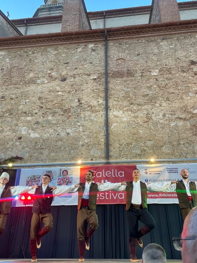 In Calella, Spain, a group of men in traditional folk costumes perform on a stage in front of a stone building. The men are wearing white shirts, brown vests, and brown pants tucked into tall, patterned socks with shoes. They are positioned in a line, with their arms extended to the sides, and each is balancing on one leg. Behind them, a banner with text that includes "Calella" and "Folklore Festival" is visible. The backdrop is a dark curtain, and the stage is lit by spotlights. The stone building behind the stage appears to be ancient, with textured walls and brick accents. The sky above is a clear blue.