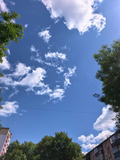 The image shows a clear blue sky with scattered white clouds, taken from a low angle looking upwards. Green trees frame the top left and right of the image, with branches extending into the frame. Two separate buildings are visible at the bottom. On the left, a tall building with white and red vertical stripes is partially shown. On the right, a brick apartment building with balconies runs along the bottom edge. Two thin black wires stretch across the sky, one higher than the other. The scene appears to be an urban environment on a sunny day. The location is the city of Sterlitamak, Russia.