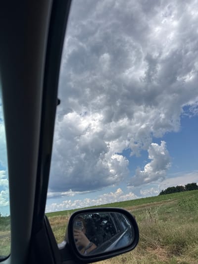 The image shows a view from inside a car, looking out towards a cloudy sky and a rural landscape. In the foreground, a portion of the car's interior is visible on the left side, and a side-view mirror on the right. The side-view mirror reflects a person holding a phone, presumably taking the photo. The landscape outside consists of a field of green crops, with a line of trees in the distance. The sky is filled with dramatic, grey clouds interspersed with patches of blue sky. The scene appears to be taken on a sunny day with approaching clouds. The location is identified as Banya, Bulgaria.