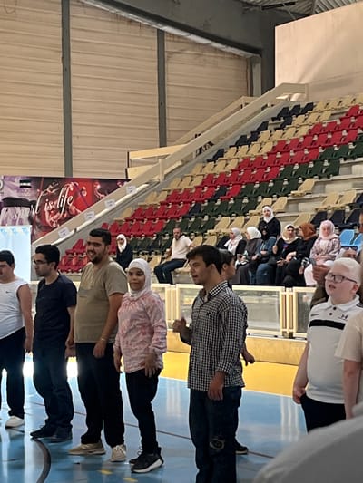 This image captures an indoor scene at what appears to be a gymnasium or event hall in Al-Qanawat Municipality, Syria. The photograph is oriented 90 degrees clockwise.

In the foreground and mid-ground, a line of approximately eight to ten individuals, mostly young to middle-aged men and one woman wearing a headscarf, are standing upright. They are arranged in a loose line, facing towards the right side of the frame, possibly participating in an exercise, training, or awaiting instruction. Most have neutral to slightly smiling expressions. They are dressed in casual attire, including shirts, t-shirts, and jeans. The floor beneath them is light blue with yellow lines, typical of a sports court.

In the background, multiple rows of tiered stadium-style seating are visible, filled with numerous spectators. The seats are arranged in a pattern of red, green, beige, and dark blue/black. Many of the people seated, particularly women, are wearing headscarves, while men are dressed casually. They appear to be observing the activity taking place on the floor.

The venue features white structural beams and railings, suggesting an interior construction. On the far left wall, a large poster or banner with stylized white Arabic script against a dark background is partially visible. The lighting is bright and artificial, typical of an indoor facility. The overall atmosphere suggests an organized event or community gathering.