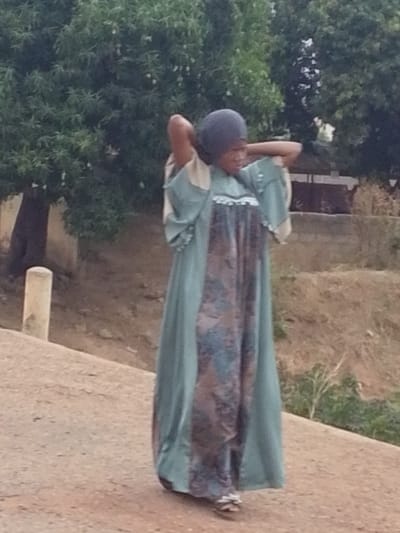 A woman in Dungu, Ghana stands on a dirt path or road. She is wearing a long, flowing garment with a green outer layer and a patterned inner layer. Her head is covered with a dark fabric. Her arms are raised behind her head. Behind her is a tree and foliage.